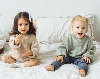 Puede incluir: Tres niños sentados juntos, vistiendo suéteres de punto de manga larga en color crema, beige y verde salvia. El niño de la izquierda lleva pantalones marrones, el del centro un pelele de punto beige y el de la derecha pantalones gris oscuro.