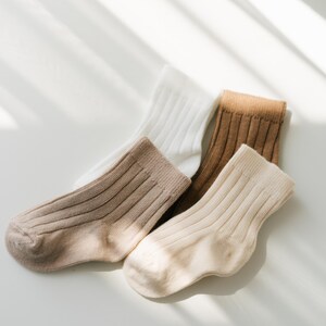 May include: Three pairs of ribbed socks in different shades of brown and white. The socks are folded and arranged on a white surface.