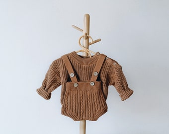 May include: A brown knitted sweater and matching overalls with brown buttons. The outfit is hanging on a wooden hanger.