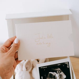 May include: A white gift box containing a pair of plush white rabbit slippers and a black and white ultrasound scan. A white card is held in front of the box with the gold embossed text "Just a little note to say..." printed on the front surface.