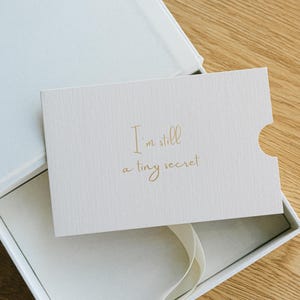 May include: A rectangular off-white card with a textured surface featuring the gold foil text "I'm still a tiny secret" in a cursive script, resting inside an open white gift box with a white ribbon pull tab visible at the bottom edge.