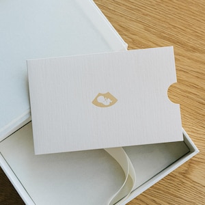 May include: A white rectangular card with a textured finish featuring a small gold-colored icon of a fetus in an ultrasound scan. The card is partially inserted into a matching white presentation box with a white ribbon pull tab visible.