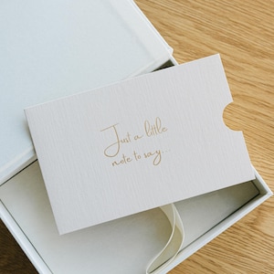 May include: A white rectangular gift box containing a textured off-white card with a thumb notch on the right side. The card features gold foil lettering that reads, "Just a little note to say..." and a white ribbon pull tab is visible.