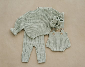 May include: A sage green knitted baby outfit set with a sweater, pants, and booties. The sweater has a round neckline and long sleeves. The pants have a cable knit pattern. The booties are soft and cozy.