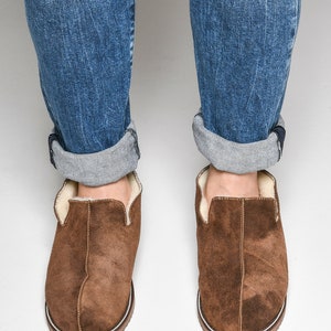 Op de afbeelding: Bruine su&egrave;de slippers met een witte rand en een voering van schapenvacht. De slippers worden gedragen door iemand die een blauwe jeans draagt.
