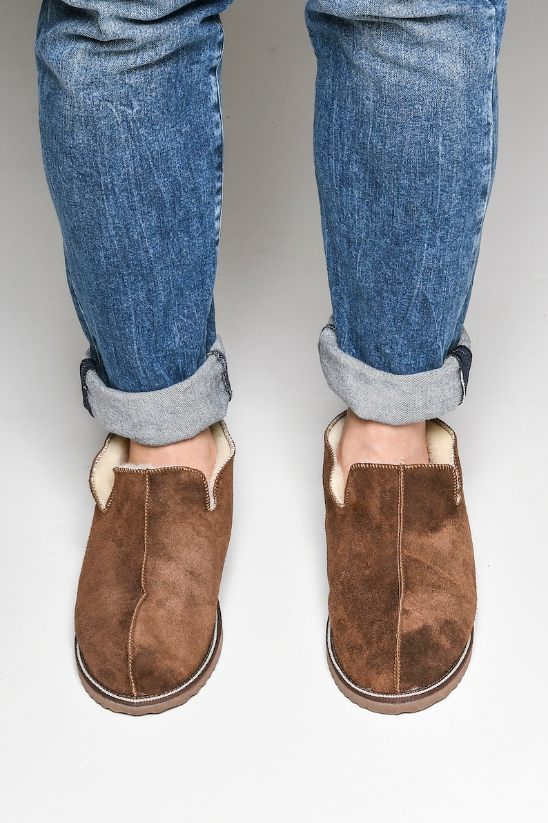 Op de afbeelding: Bruine su&egrave;de slippers met een witte rand en een voering van schapenvacht. De slippers worden gedragen door iemand die een blauwe jeans draagt.