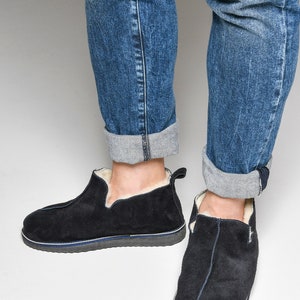 May include: A pair of black suede slippers with a white shearling lining. The slippers have a navy blue trim around the sole and a slightly pointed toe.
