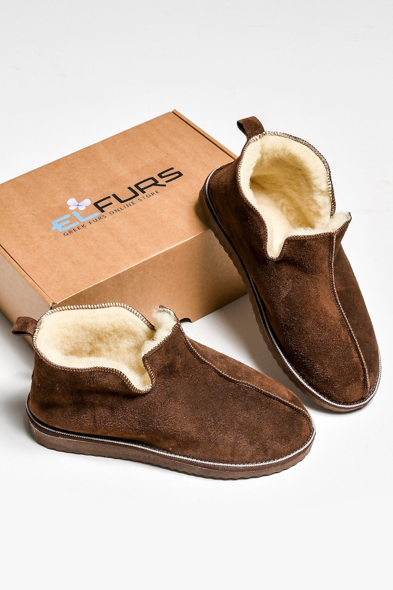 Op de afbeelding: Een paar bruine su&egrave;de pantoffels met een schapenvacht voering. De pantoffels liggen op een witte achtergrond en een bruine doos met de tekst "EL FURS GREEK FURS ONLINE STORE" is zichtbaar op de achtergrond.