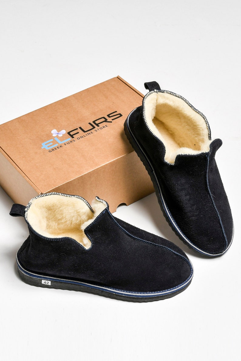 May include: A pair of black suede slippers with a white shearling lining. The slippers have a navy blue trim and a white sole. The slippers are sitting on a brown box with the text "EL FURS GREEK FURS ONLINE STORE" printed on it.