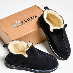 Op de afbeelding: Een paar zwarte su&egrave;de pantoffels met een witte rand en een pluche witte voering. De pantoffels liggen op een wit oppervlak en een bruine doos met de tekst "EL FUR'S GREEK FURS ONLINE STORE" is op de achtergrond.