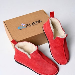 Op de afbeelding: Twee paar rode su&egrave;de pantoffels met een witte fleece voering. De pantoffels liggen op een bruine kartonnen doos met de tekst "EL FURS GREEK FURS ONLINE STORE" gedrukt op de bovenkant.