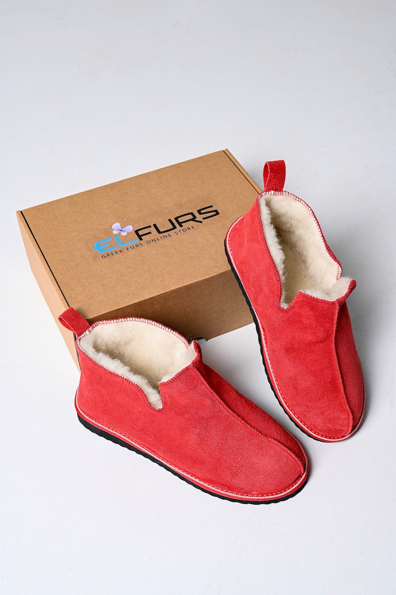 Op de afbeelding: Twee paar rode su&egrave;de pantoffels met een witte fleece voering. De pantoffels liggen op een bruine kartonnen doos met de tekst "EL FURS GREEK FURS ONLINE STORE" gedrukt op de bovenkant.