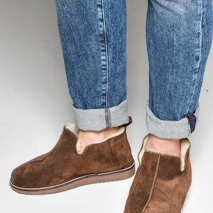 Op de afbeelding: Bruine su&egrave;de slippers met een voering van schapenvacht. De slippers hebben een afgeronde neus en een platte zool.