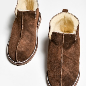 Op de afbeelding: Een paar bruine su&egrave;de pantoffels met een witte rand rond de zool. De pantoffels zijn gevoerd met een lichtbruin fleece.