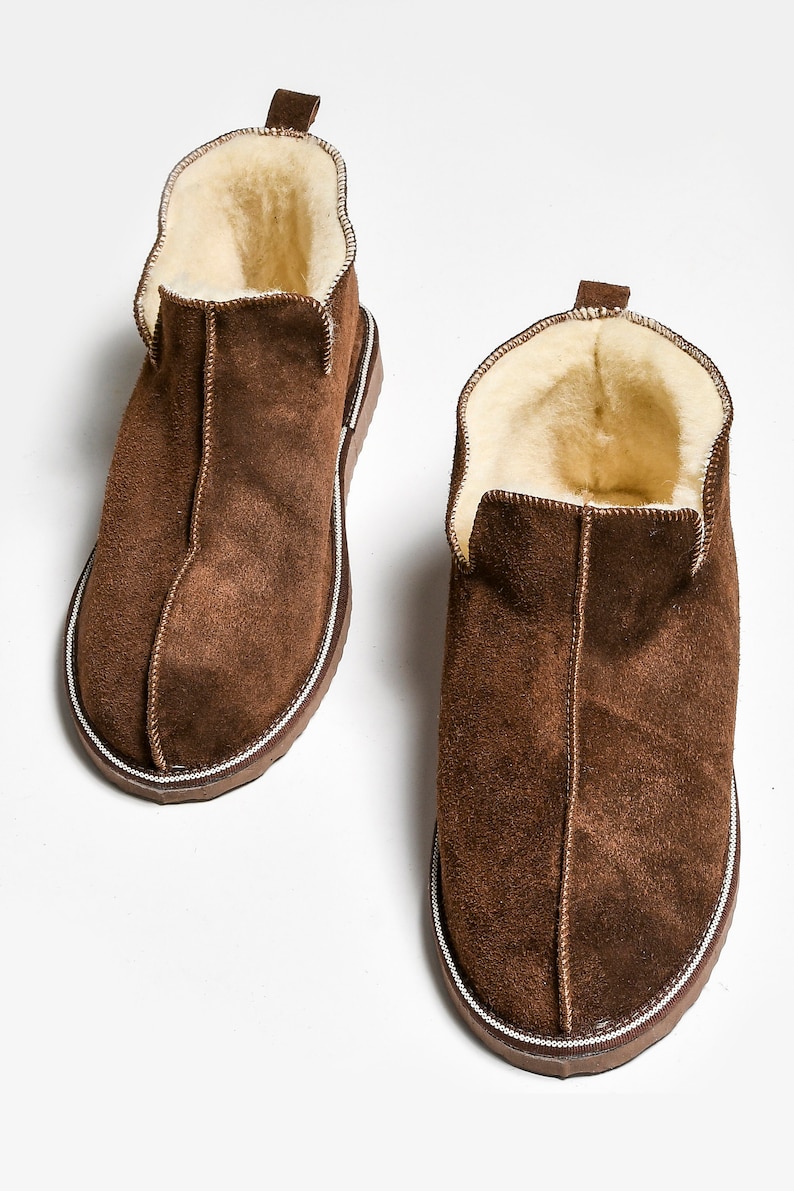 Op de afbeelding: Een paar bruine su&egrave;de pantoffels met een witte rand rond de zool. De pantoffels zijn gevoerd met een lichtbruin fleece.