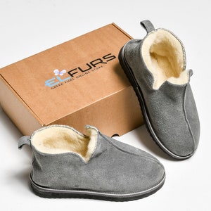 Op de afbeelding: Een paar grijze su&egrave;de pantoffels met een fleece voering. De pantoffels liggen op een wit oppervlak naast een bruine doos met de tekst "EL FURS GREEK FURS ONLINE STORE".