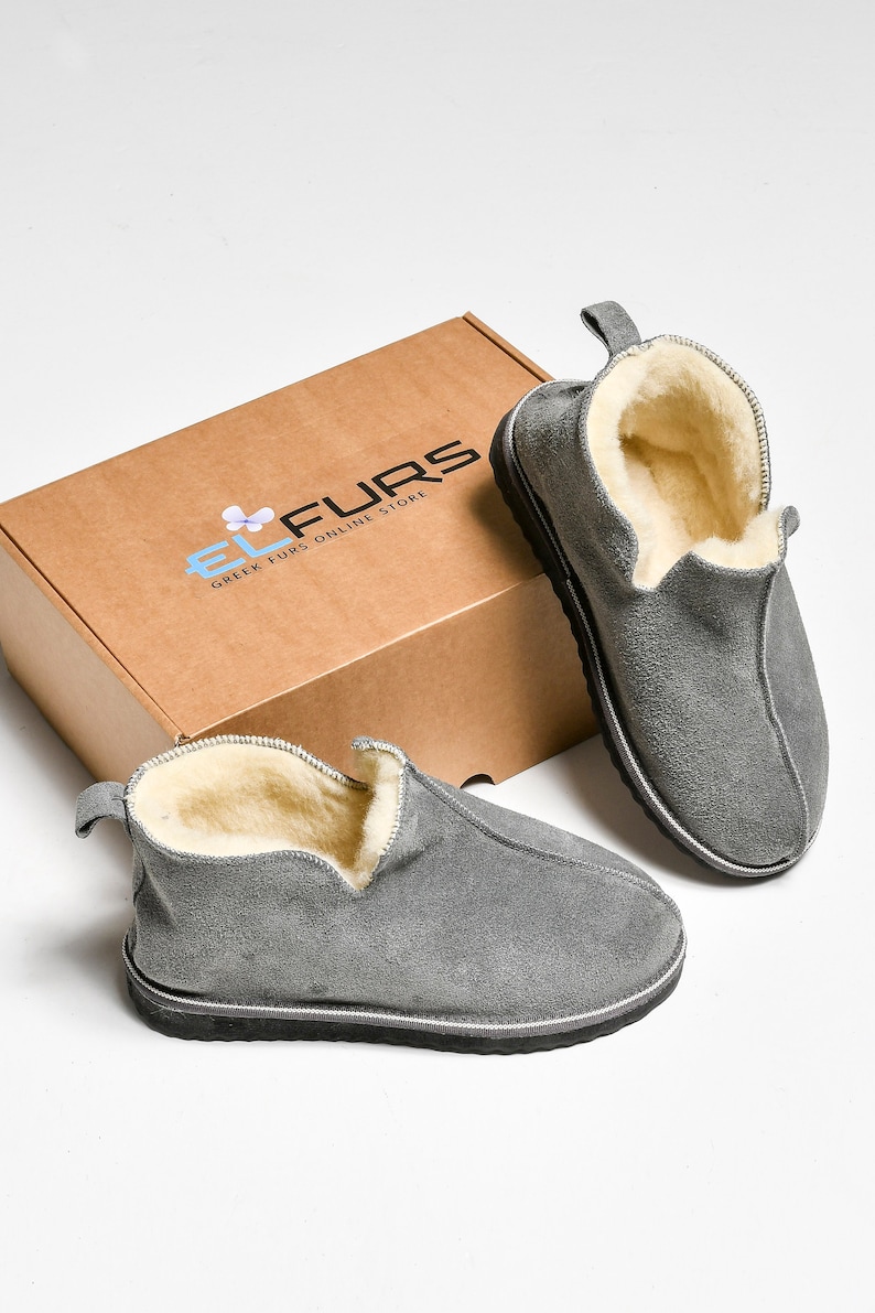 Op de afbeelding: Een paar grijze su&egrave;de pantoffels met een fleece voering. De pantoffels liggen op een wit oppervlak naast een bruine doos met de tekst "EL FURS GREEK FURS ONLINE STORE".