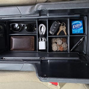 May include: A black plastic car centre console organiser tray with multiple compartments containing black sunglasses, a brown leather wallet, a white charging cable, a car key fob with keys, a pack of Extra Peppermint gum, coins, and a black pen.