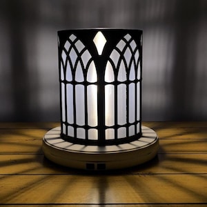 Könnte beinhalten: Eine schwarze Metalllampe mit einem weißen, mattierten Glasschirm. Der Schirm hat ein gotisches Bogenmuster. Die Lampe steht auf einem weißen Sockel mit einem Stromkabel.