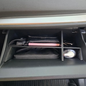 Puede incluir: Guantera de coche abierta con varios objetos organizados en su interior. Se ven una cartera de cuero negro, un cuaderno rosa, gafas de sol y un bote de gel desinfectante. El compartimento está dividido en secciones.