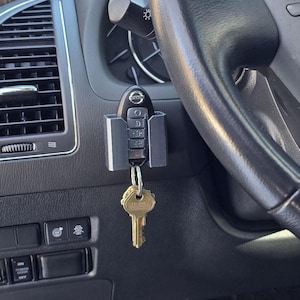 May include: A silver car key holder with a black key fob attached. The key holder is mounted on the dashboard of a car. The key fob has the Nissan logo and buttons.