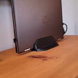 May include: A dark gray laptop computer with the Dell logo is propped up on a black stand. The laptop is on a wooden surface. A monitor is in the background.