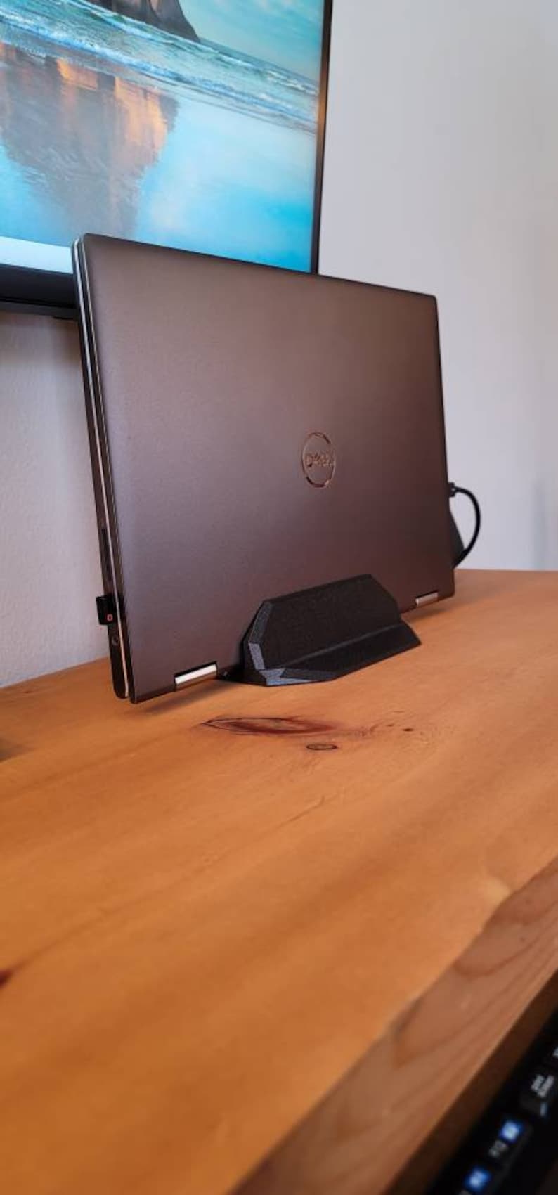 May include: A dark gray laptop computer with the Dell logo is propped up on a black stand. The laptop is on a wooden surface. A monitor is in the background.