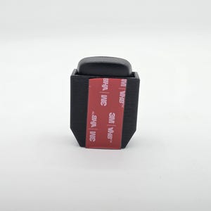 May include: A black, rectangular object with a rounded top. The object has a red and white 3M VHB adhesive strip on the front. The object is likely a mounting bracket or holder.
