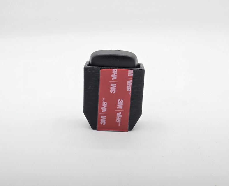 May include: A black, rectangular object with a rounded top. The object has a red and white 3M VHB adhesive strip on the front. The object is likely a mounting bracket or holder.
