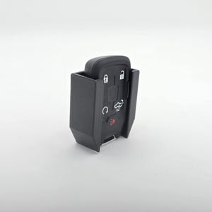 May include: A black key fob with several buttons and a red light, encased in a black holder. The key fob has symbols for lock, unlock, and other functions. The holder appears to be 3D printed.