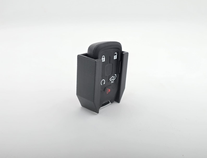 May include: A black key fob with several buttons and a red light, encased in a black holder. The key fob has symbols for lock, unlock, and other functions. The holder appears to be 3D printed.