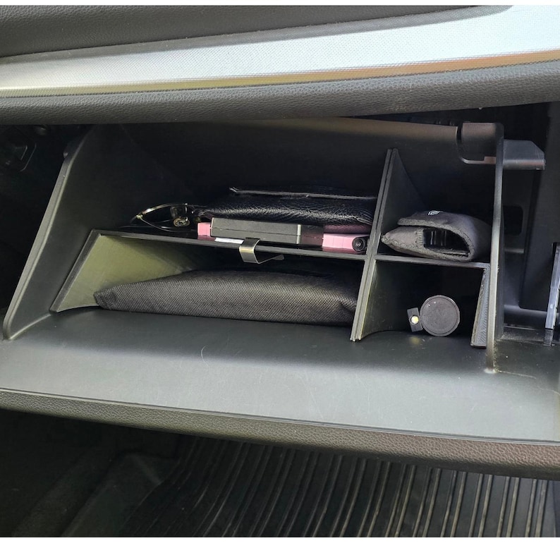 K&ouml;nnte beinhalten: Ein offenes Handschuhfach im Auto, das eine organisierte Aufbewahrung zeigt. Im Inneren sind eine schwarze Geldb&ouml;rse, ein pinkfarbenes Telefon, ein schwarzer Beutel und ein kleiner, runder schwarzer Gegenstand zu sehen. Das Fach besteht aus schwarzem Kunststoff.