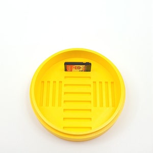 May include: A yellow plastic container with a grid pattern on the inside. A game cartridge is sitting inside the container.