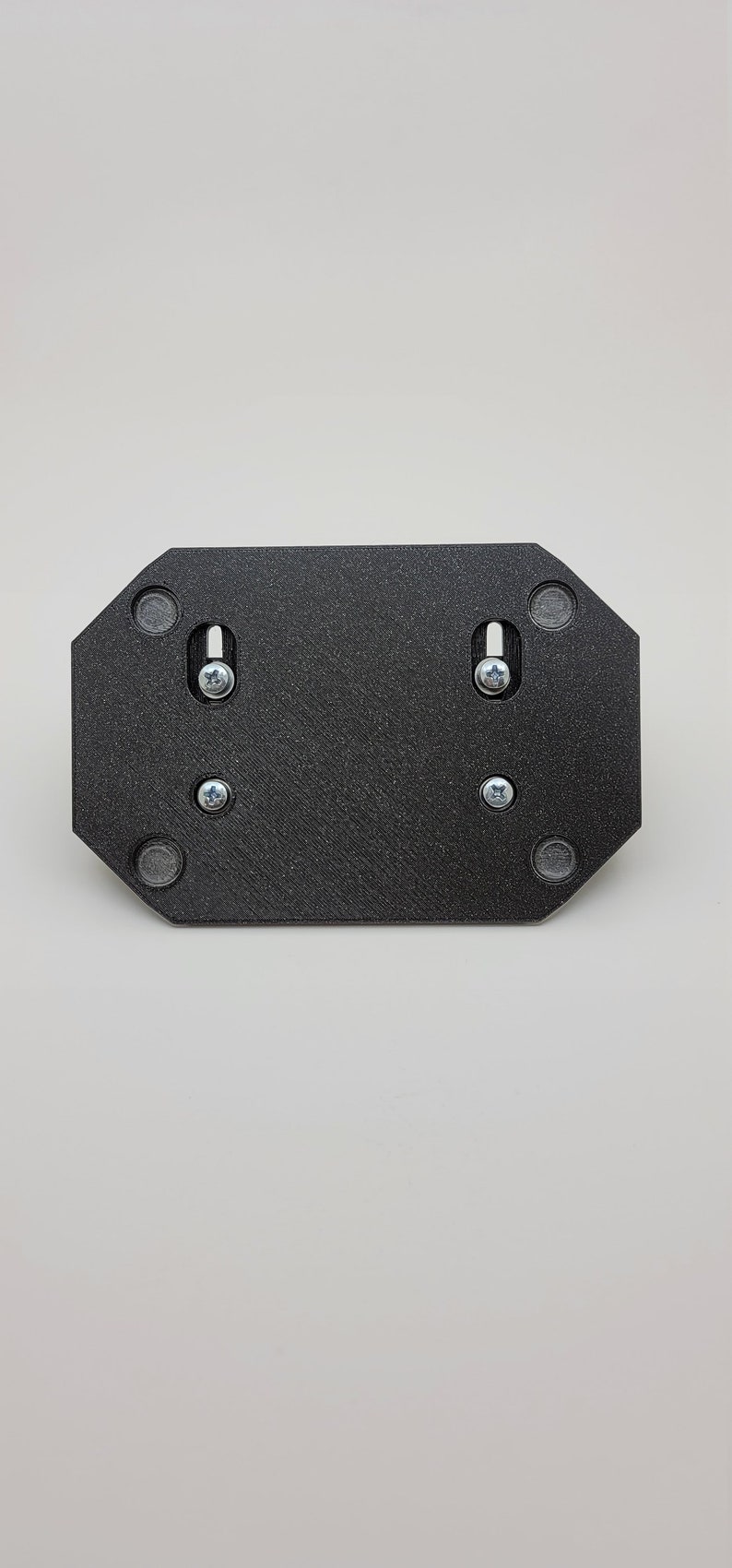 May include: A black, octagonal mounting plate with a textured surface. It features two screws and four small, circular magnets. The plate is designed for mounting and is shown against a white background.