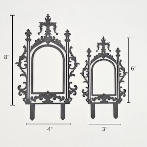 May include: Two black decorative garden stakes. The larger stake is 20 cm tall and 10 cm wide, while the smaller is 15 cm tall and 8 cm wide. Both have an arched window design with ornate detailing and a stake at the bottom.