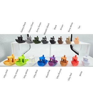 May include: Two rows of small toy boats in various colors, including gold, green, white, blue, pink, bronze, red, and tan. The boats are arranged in a row on a white surface.