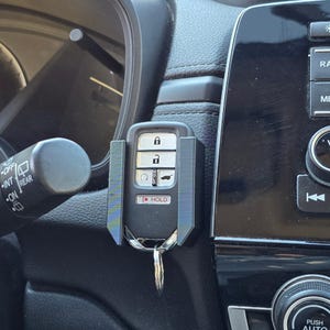 May include: A black car key with a key holder attached to a car dashboard. The key has buttons for locking, unlocking, and other functions. The key holder is black and holds the key securely in place.