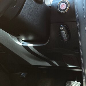 May include: Close-up of a black car interior with a key fob and a start/stop button. The key fob has a silver Nissan logo and the start/stop button has a red circle with the words "Start Stop" in white.