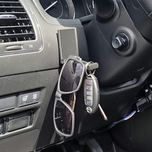 May include: A black car interior with a black sunglasses holder attached to the dashboard. The holder has a pair of black sunglasses and a car key attached to it.