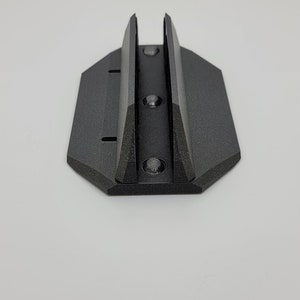 May include: A black, 3D-printed mounting bracket with a textured surface. The bracket has a rectangular base with a hexagonal shape and two vertical supports. Three screw holes are visible.
