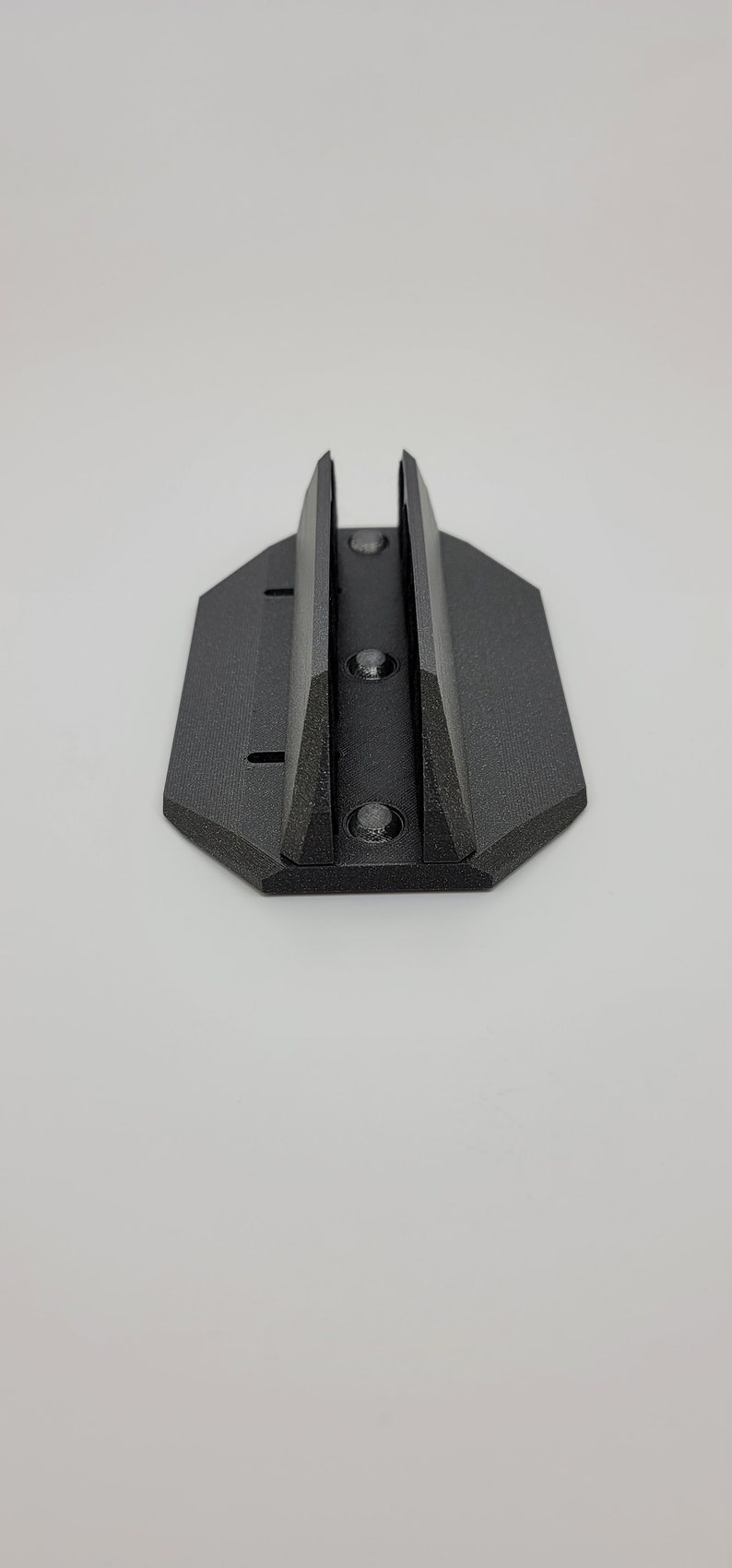 May include: A black, 3D-printed mounting bracket with a textured surface. The bracket has a rectangular base with a hexagonal shape and two vertical supports. Three screw holes are visible.