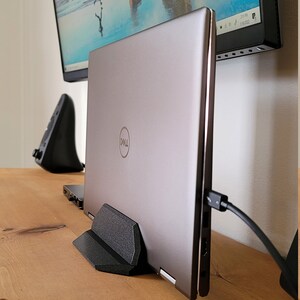 May include: A silver Dell laptop is propped up on a black stand on a wooden desk. A monitor displaying a beach scene is mounted on the wall behind the laptop. A black mouse and a black cable are also visible.