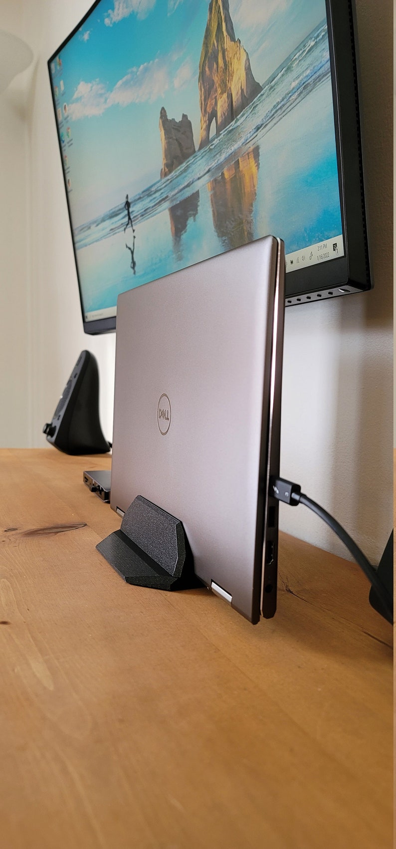 May include: A silver Dell laptop is propped up on a black stand on a wooden desk. A monitor displaying a beach scene is mounted on the wall behind the laptop. A black mouse and a black cable are also visible.