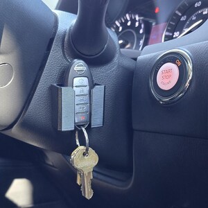 May include: A black car interior with a key fob attached to a key holder. The key fob has a silver NISSAN logo and buttons. The car has a red START STOP ENGINE button.