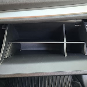 K&ouml;nnte beinhalten: Ge&ouml;ffnete schwarze Auto-Handschuhfach mit silberner Zierleiste. Das Fach ist in Abschnitte mit einem Regal unterteilt. Der Innenraum ist schwarz mit einer strukturierten Oberfl&auml;che.