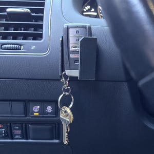 May include: A black car key fob and keys attached to a car dashboard. The key fob is held in a black holder. The keys are attached to the fob with a silver keyring. The dashboard is dark grey with air vents.
