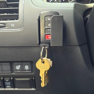 May include: A black key fob holder attached to a car's dashboard, holding a key fob and a set of gold keys. The key fob has buttons for locking, unlocking, and other functions. The keys are attached to a metal ring.