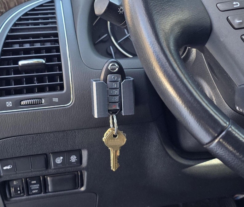 May include: A silver car key holder with a black key fob attached. The key holder is mounted on the dashboard of a car. The key fob has the Nissan logo and buttons.