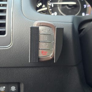 May include: A car key fob is held in a black 3D-printed holder, mounted on a car's dashboard. The key fob has several buttons with icons and the word "HOLD" in red. The dashboard is gray.