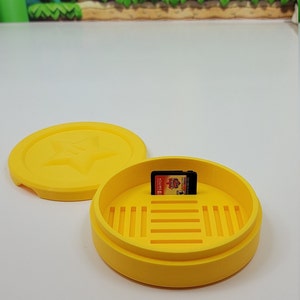 May include: Yellow plastic case with a lid and a game cartridge inside. The case has a star design on the lid and a grid pattern on the bottom. The game cartridge is for the Nintendo Switch.